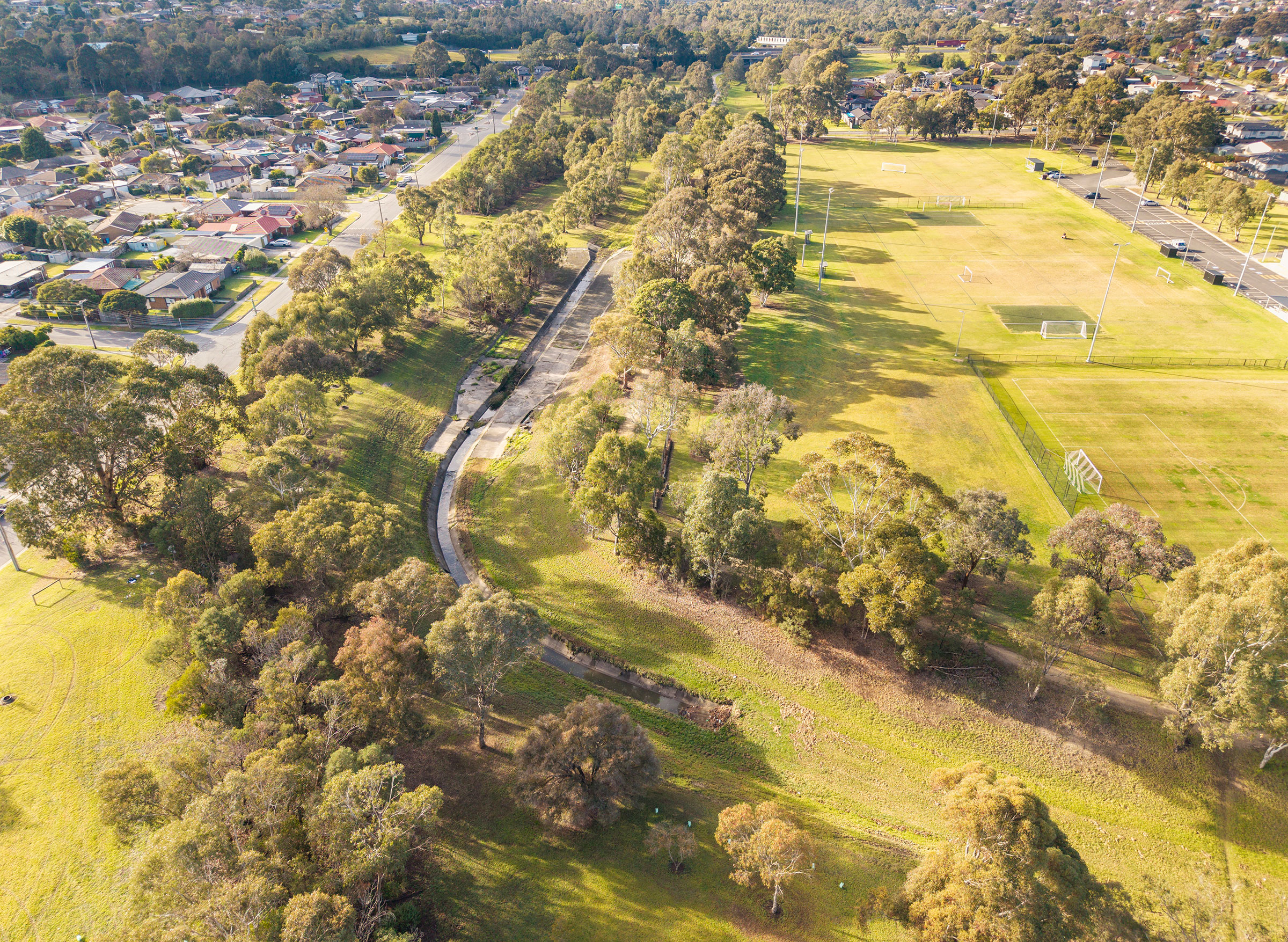 Aerial view of a concreted section of Eumemmerring Creek running through a grassy area surrounded by houses and sporting facilities.