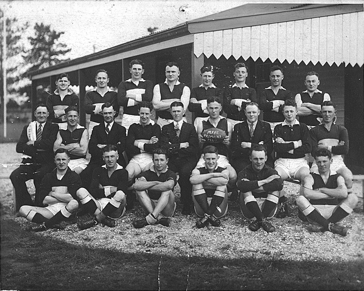 Black and white photo of group of men in football jumpers outside a wooden pavilion