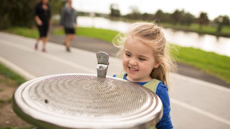 Young girl stands beside public drinking water fountain with river in the background
