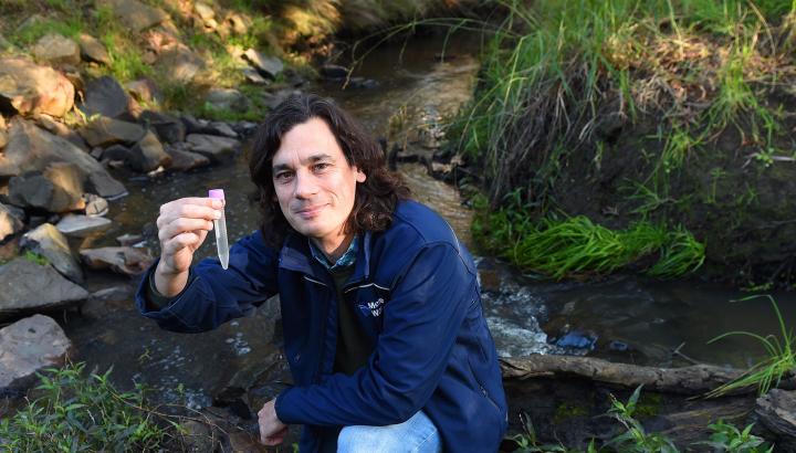 Rhys Coleman crouches by a waterway, holding a test tube.