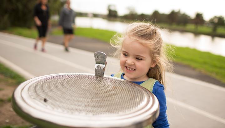 Young girl stands beside public drinking water fountain with river in the background