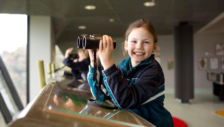A young girl is smiling at the camera with the Edithvale Wetlands lookout in the background. She is holding a pair of binoculars.