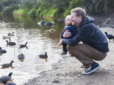 Man and baby sit by waterway surrounded by birds