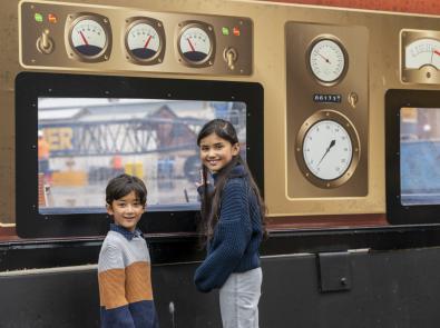 Two children in front of construction hoarding for the Hobsons Bay Main Sewer. The hoarding is decorated with old-fashioned gauges, and the construction site is visible through clear cut-out windows.