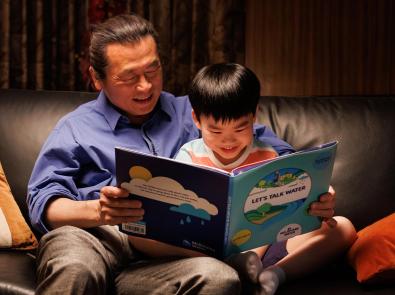 Father and young son sit on a couch reading a picture book, titled "Let's talk water"