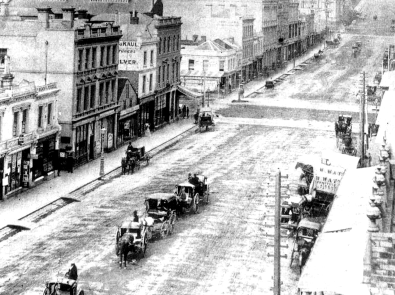 Black and white photograph of horse and carriages crossing wide street with drain running alongside