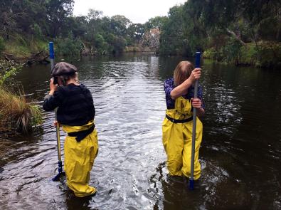 Two people in waders stand in a river, holding long poles to collect water samples
