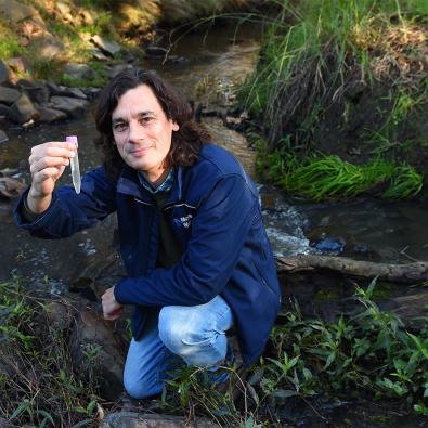 Research scientist Rhys Coleman crouches by a waterway, holding a test tube.