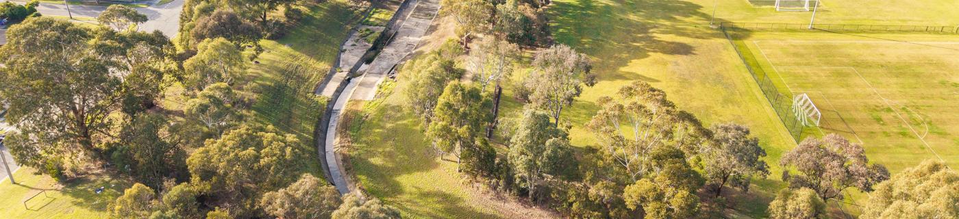 Aerial view of a concreted section of Eumemmerring Creek running through a grassy area surrounded by houses and sporting facilities.