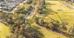 Aerial view of a concreted section of Eumemmerring Creek running through a grassy area surrounded by houses and sporting facilities.