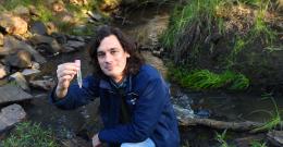 Rhys Coleman crouches by a waterway, holding a test tube.