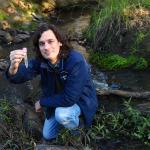 Research scientist Rhys Coleman crouches by a waterway, holding a test tube.