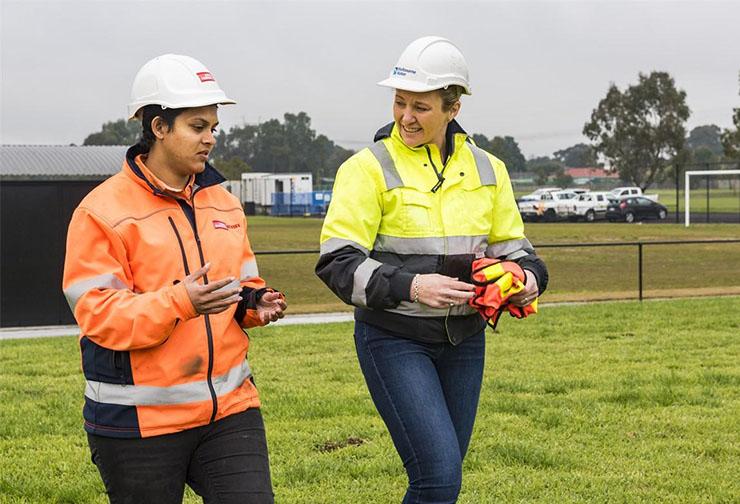 Two women in hard hats and high-visibility workwear talk at a project site