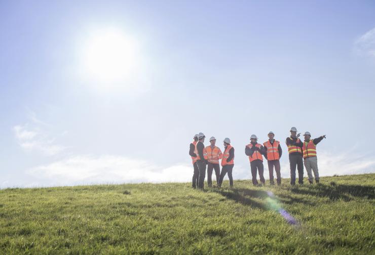 Group of men in high-vis and hard hats standing on a green hill with the sun behind them