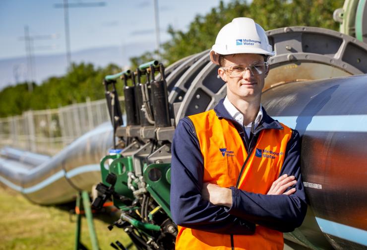 White man in Melbourne Water PPE leaning against large pipe, arms crossed and looking directly into camera