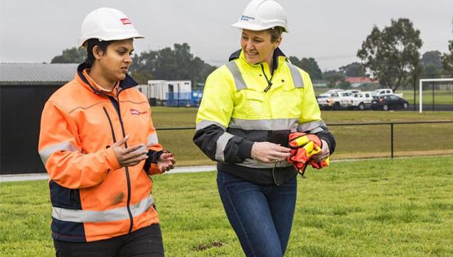 Two women in hard hats and high-visibility workwear talk at a project site
