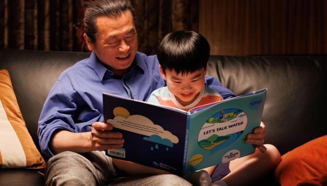 Father and young son sit on a couch reading a picture book, titled "Let's talk water"