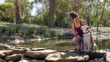 Woman in exercise gear kneels next to her dog on a path across a creek