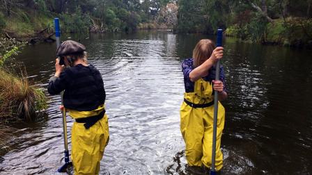 Two people in waders stand in a river, holding long poles to collect water samples